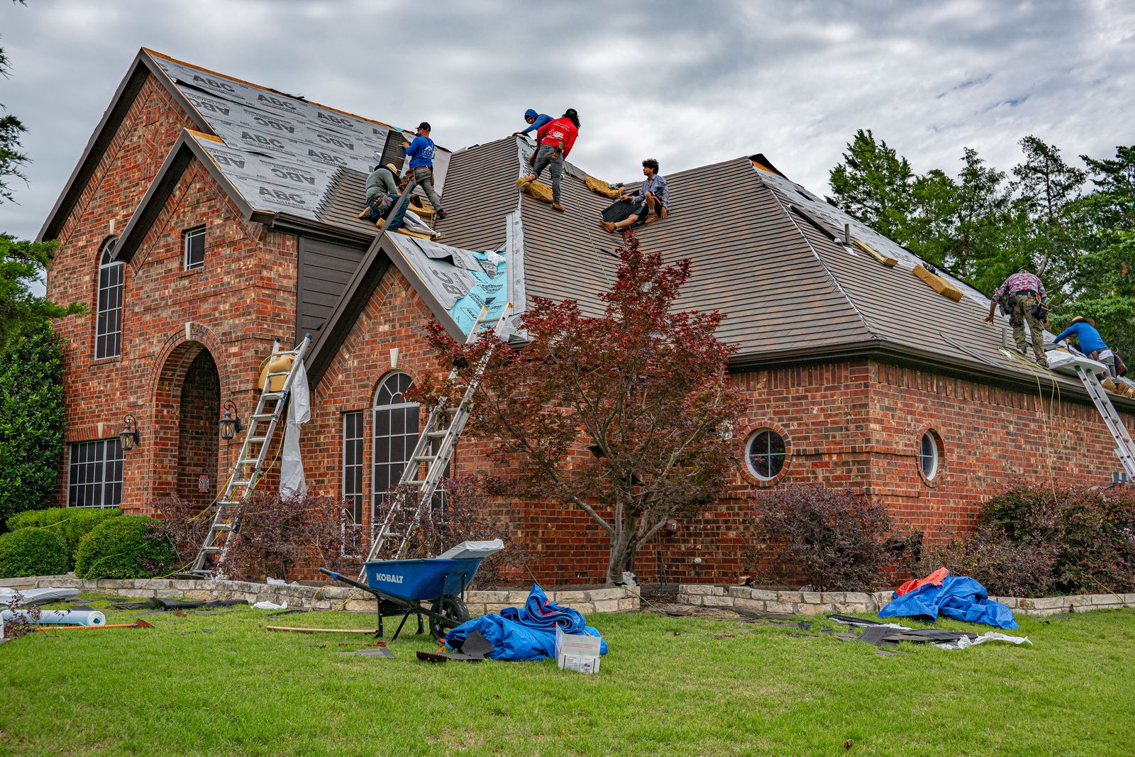 Roofing crew replacing the shingle roof on a brick home, ladders against the eaves